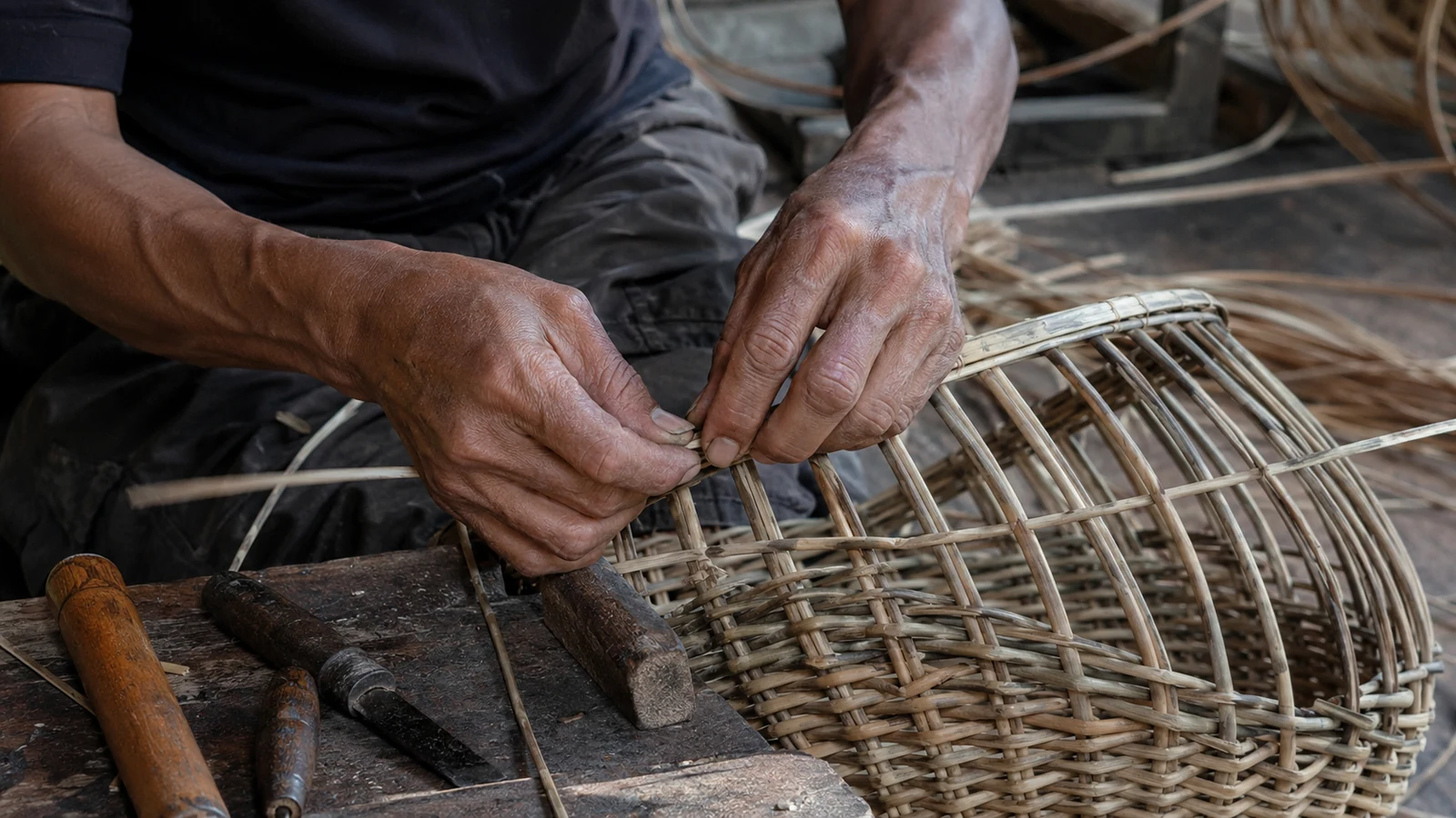 An artisan weaving natural rattan strips around a basket frame in a workshop.